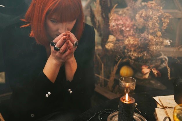 Beautiful red haired young woman in black hat and witch outfit does magic in greenhouse decorated for Halloween celebration with pumpkins and skull in the smoke