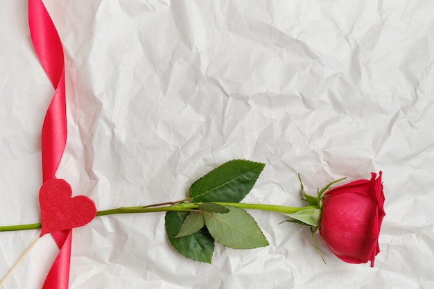 Beautiful red rose with ribbon and heart on a stick in the background on crumpled paper. Top view. Copy space.