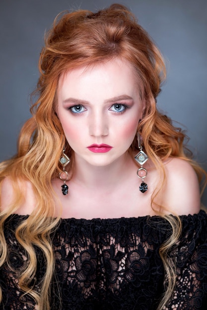 A beautiful redhaired girl with wavy hair makeup bare shoulders and long earrings posing on a gray background in the studio woman looking at camera