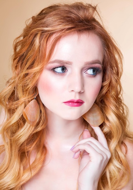 A beautiful redhaired girl with wavy hair and makeup poses on a beige background in the studio in round gold earrings Woman touching an earring and looking away