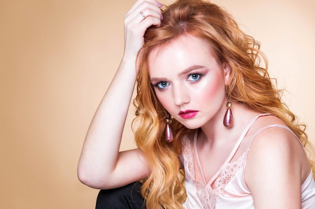 Beautiful redhaired girl with wavy hair makeup in a silk top and long earrings posing on a beige background in the studio woman looking at camera