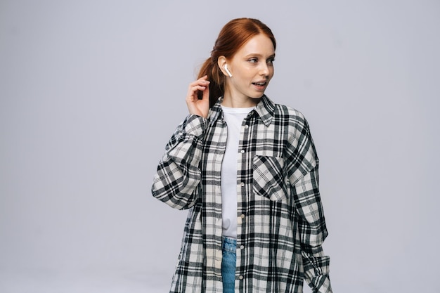 Beautiful redhaired young woman wearing wireless earphones listening popular song on isolated white background looking away Pretty redhead lady model emotionally showing facial expressions