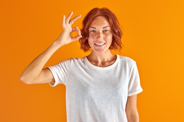 Beautiful redhead woman doing ok sign with hand and fingers isolated over yellow space