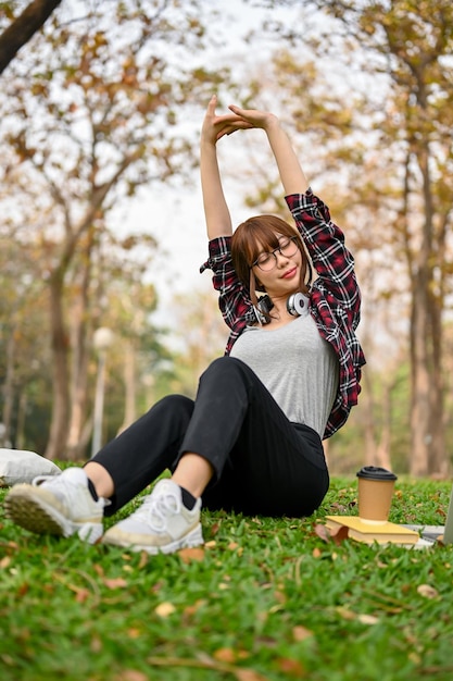 Beautiful and relaxed young Asian female stretching her hands sitting on grass in the park