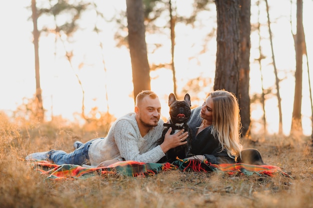 Beautiful romantic couple is having fun with their dog french bulldog on outdoors in the autumn forest at sunset