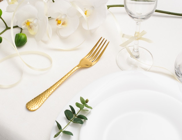 Beautiful romantic table setting with gold cutlery, orchid and white empty plate on white background