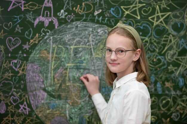 A beautiful schoolgirl in glasses stands at a painted blackboard and looks into the camera