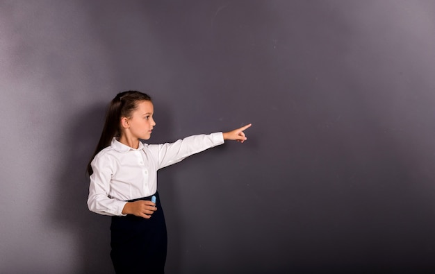 A beautiful schoolgirl in a uniform stands and points with her hand on a black background with a place for text