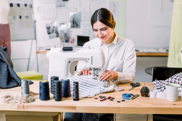 Beautiful seamstress sewing at her workshop. Dressmaker working on the sewing machine. Tailor making a garment in her workplace.