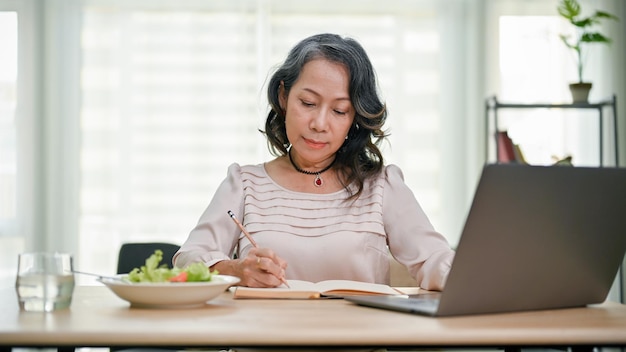 Beautiful senior asian businesswoman working from home focused on writing at her desk