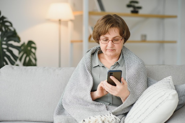 Beautiful senior woman in casual clothing using her smart phone while sitting on the sofa at home