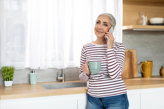 Beautiful Senior Woman Talking On Mobile Phone And Drinking Tea In Kitchen