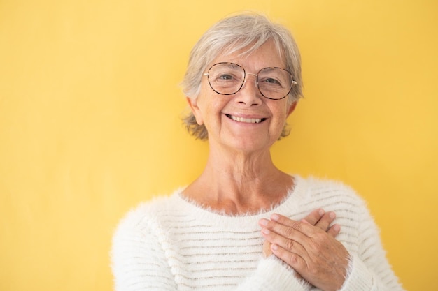 Beautiful senior woman with eyeglasses looking at camera with hands over her heart expressing love Isolated on yellow background