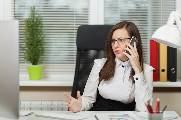 Beautiful serious business woman in suit and glasses sitting at the desk, working at contemporary computer with documents in light office, talking on mobile phone resolving issues, looking aside