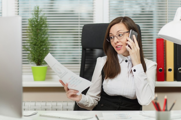 Beautiful serious business woman in suit and glasses sitting at the desk, working at contemporary computer with documents in light office, talking on mobile phone resolving issues, looking aside