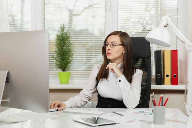 The beautiful serious and engrossed brown-hair business woman in suit and glasses sitting at the desk with tablet, working at computer with modern monitor with documents in light office, looking aside