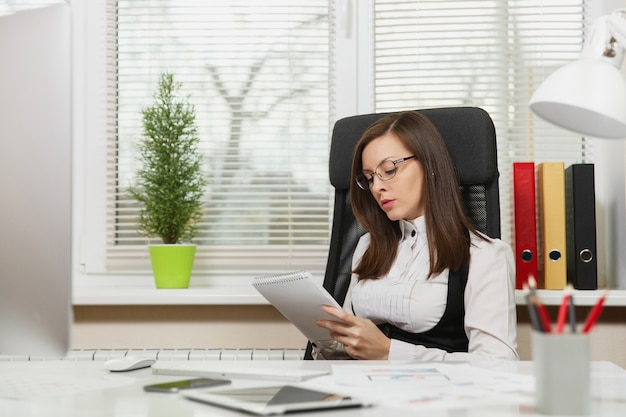 The beautiful serious and engrossed brown-hair business woman in suit and glasses sitting at the desk with tablet, working at computer with modern monitor with documents in light office, looking aside