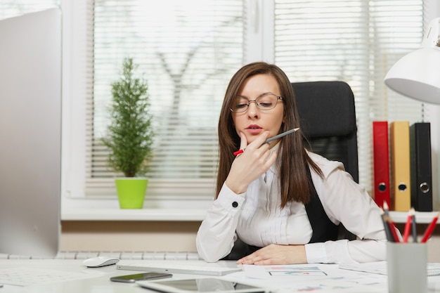 The beautiful serious and engrossed brown-hair business woman in suit and glasses sitting at the desk with tablet, working at computer with modern monitor with documents in light office, looking aside