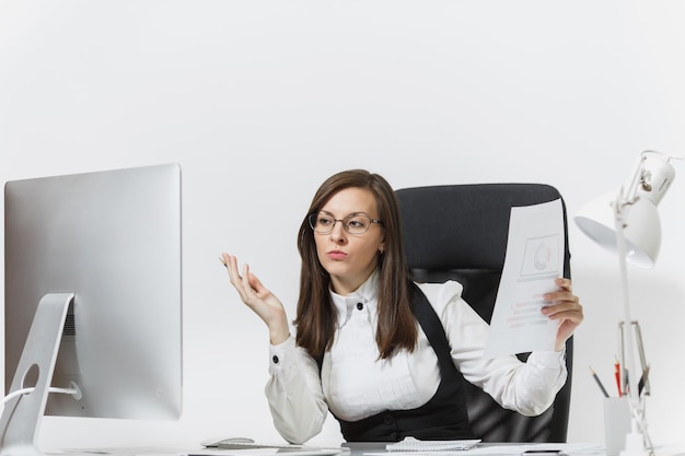 The beautiful serious and engrossed brown-hair business woman in suit and glasses sitting at the desk, working at computer with modern monitor with documents in light office,