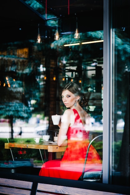 Beautiful serious European brunette girl in an evening red dress sitting at the window with a cup of coffee at the cafe.