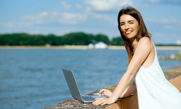 Beautiful, serious girl in a white dress with a tablet, pen, laptop phone on the dock. High quality photo