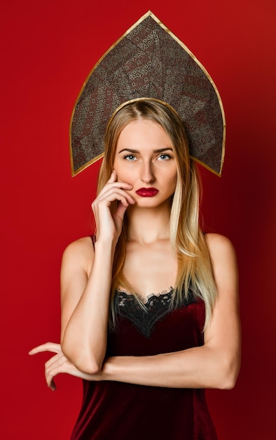 Beautiful sexy blonde Russian girl in traditional kokoshnik hat velvet festive dress on a red background