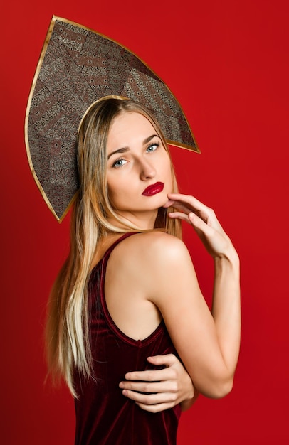 Beautiful sexy blonde Russian girl in traditional kokoshnik hat velvet festive dress on a red background