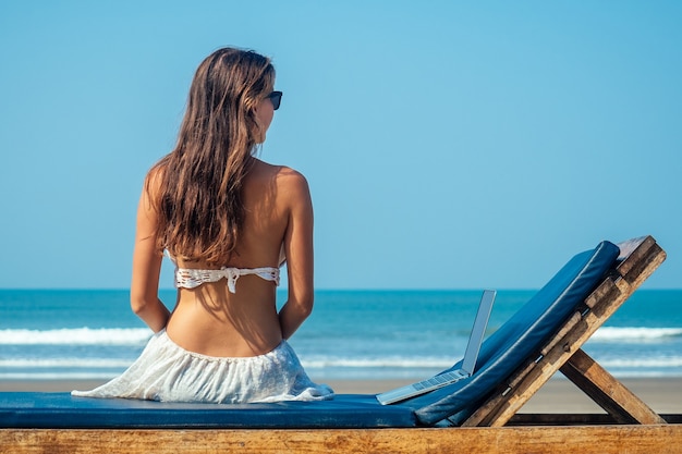 Beautiful and sexy young woman sitting on a lounger with a laptop by the sea. woman freelancer in the resort.