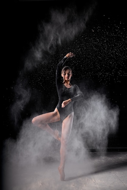 Photo beautiful slender ballet dancer women wearing black bodysuit, sensually poses among flying flour which covers her body, on black studio background. artistic, commercial, monochrome design.