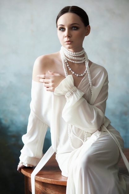 Beautiful slender brunette girl sitting on sofa in long white dress. Portrait woman with jewellery on neck