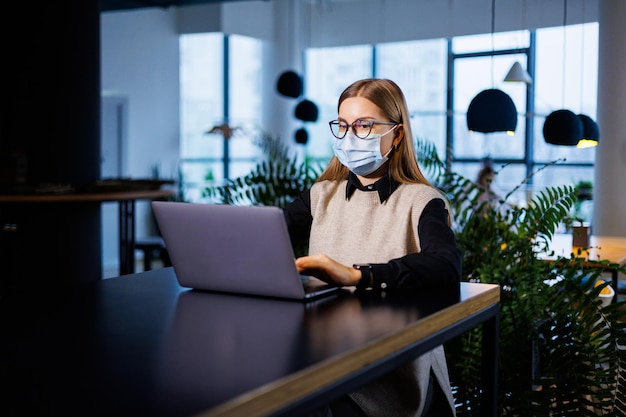 A beautiful smart businesswoman in a large company holds an online meeting with business partners in a protective mask during the coronavirus, she sits at a table at a workplace with a laptop