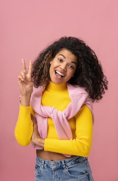 Beautiful smiling African American woman showing victory sign looking at camera isolated on pink