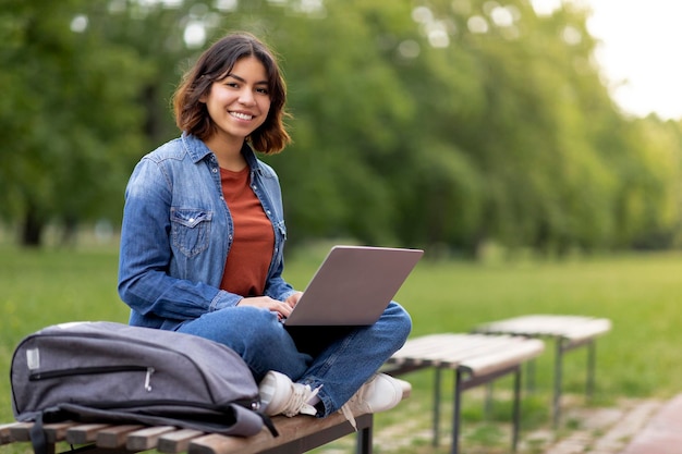 Beautiful smiling arab female student using laptop while relaxing on bench outdoors