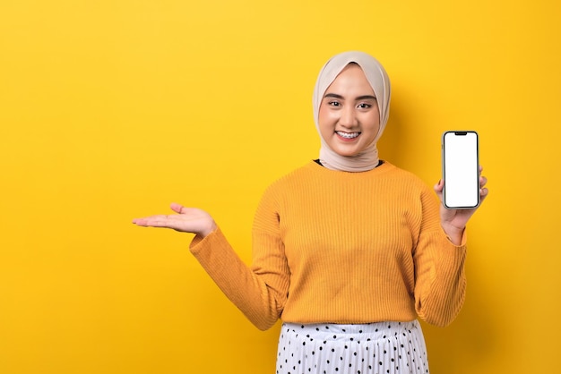 Beautiful smiling Asian girl wearing hijab holding mobile phone with blank white screen demonstrating empty space on palm isolated over yellow background mockup copy space