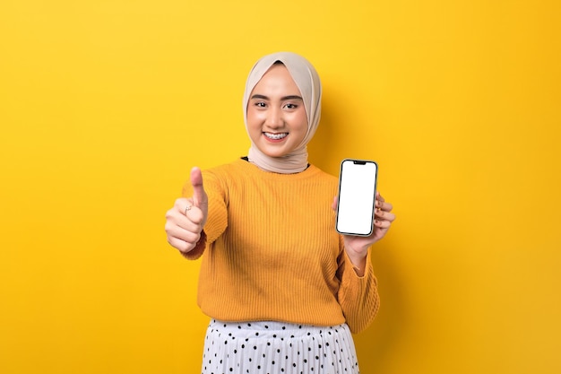 Beautiful smiling Asian girl wearing hijab holding mobile phone with blank white screen giving thumbs up isolated on yellow background mockup copy space