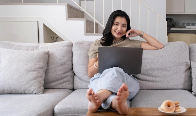 Beautiful smiling Asian woman looking at camera relaxing on leather sofa at home work on a laptop computer