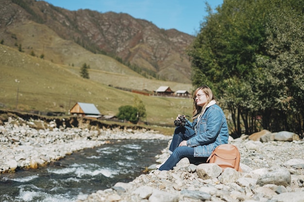 Beautiful smiling blonde young woman traveler with photo camera in hands near the mountain river