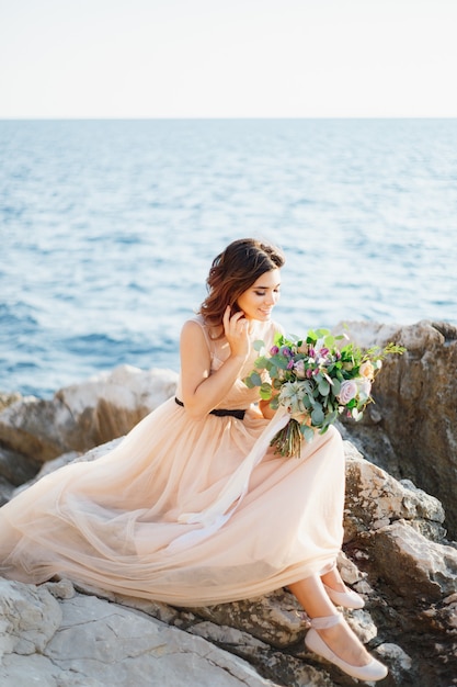 Beautiful smiling bride in a beige dress with a bouquet of flowers in her hands sits on a rock above