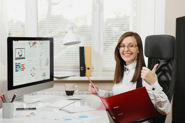 Beautiful smiling brown-hair business woman in black white suit, sitting at the desk with red folder, mobile phone, cup of coffee, showing thumb up, working at computer with documents in light office.