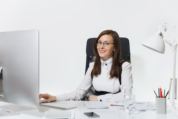 Beautiful smiling brown-hair business woman in suit and glasses sitting at the desk with glass of pure water, working at computer with modern monitor with documents in light office