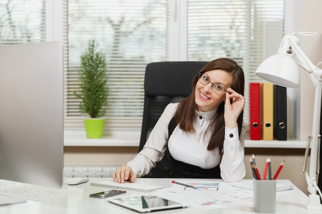 The beautiful smiling brown-hair business woman in suit and glasses sitting at the desk with tablet, working at computer with modern monitor with documents in light office, looking at the camera