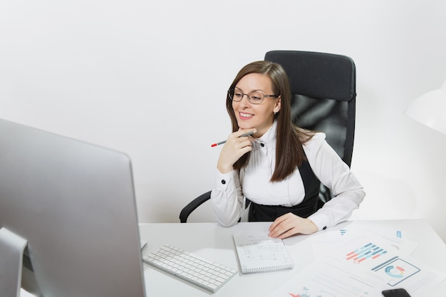 Beautiful smiling brown-hair business woman in suit and glasses sitting at the desk, working at computer with contemporary monitor with documents in light office, looking aside