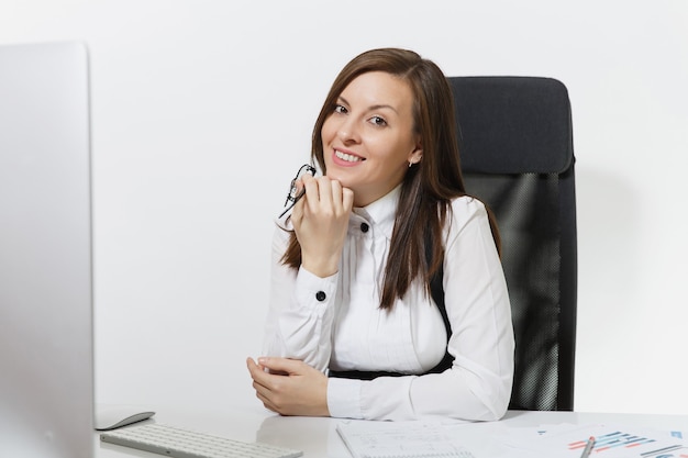 Beautiful smiling brown-hair business woman in suit and glasses sitting at the desk, working at computer with modern monitor with documents in light office.