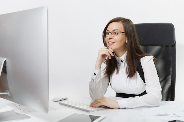 Beautiful smiling brown-hair business woman in suit and glasses sitting at the desk, working at computer with modern monitor with documents in light office.