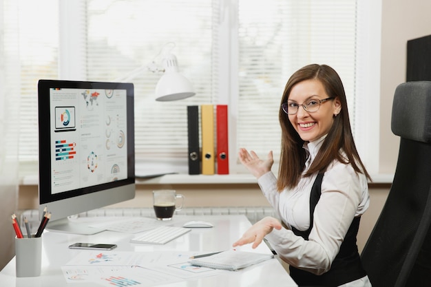 The beautiful smiling brown-hair business woman in suit and glasses working at computer with documents in light office, looking at the camera