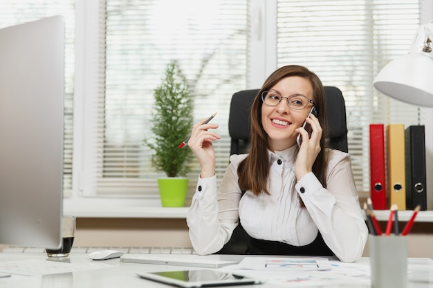 Beautiful smiling business woman in suit and glasses sitting at the desk, working at contemporary computer with documents in light office, talking on mobile phone, conducting pleasant conversation.