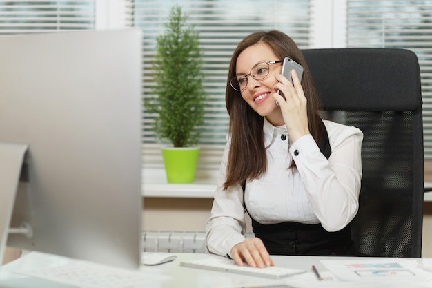 Beautiful smiling business woman in suit and glasses sitting at the desk, working at contemporary computer with documents in light office, talking on mobile phone, conducting pleasant conversation