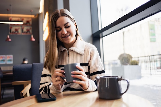 Beautiful smiling calm young woman drinking coffee looking out the window while sitting at a table in a restaurant. Morning coffee