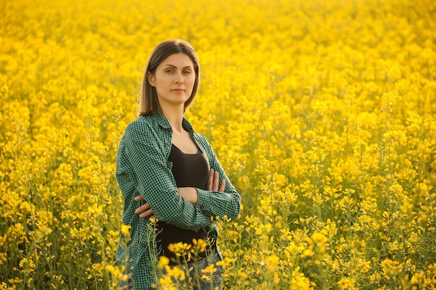 Beautiful smiling farmer woman standing yellow flowering rapeseed crossed her arms and looking at ca