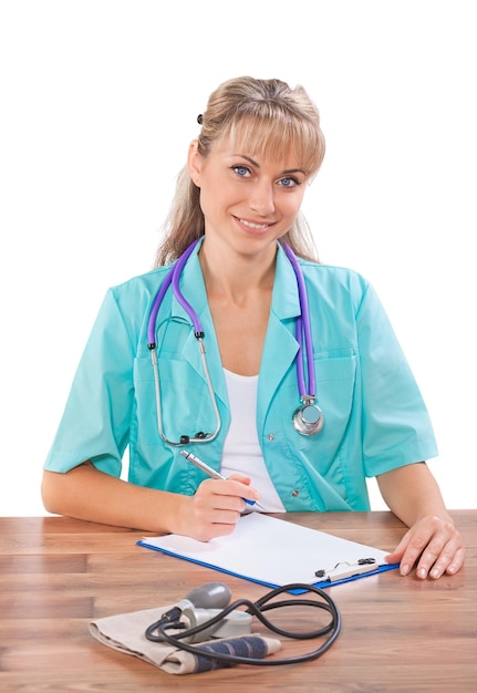 Beautiful smiling female doctor sitting at the table writing and looking at camera isolated